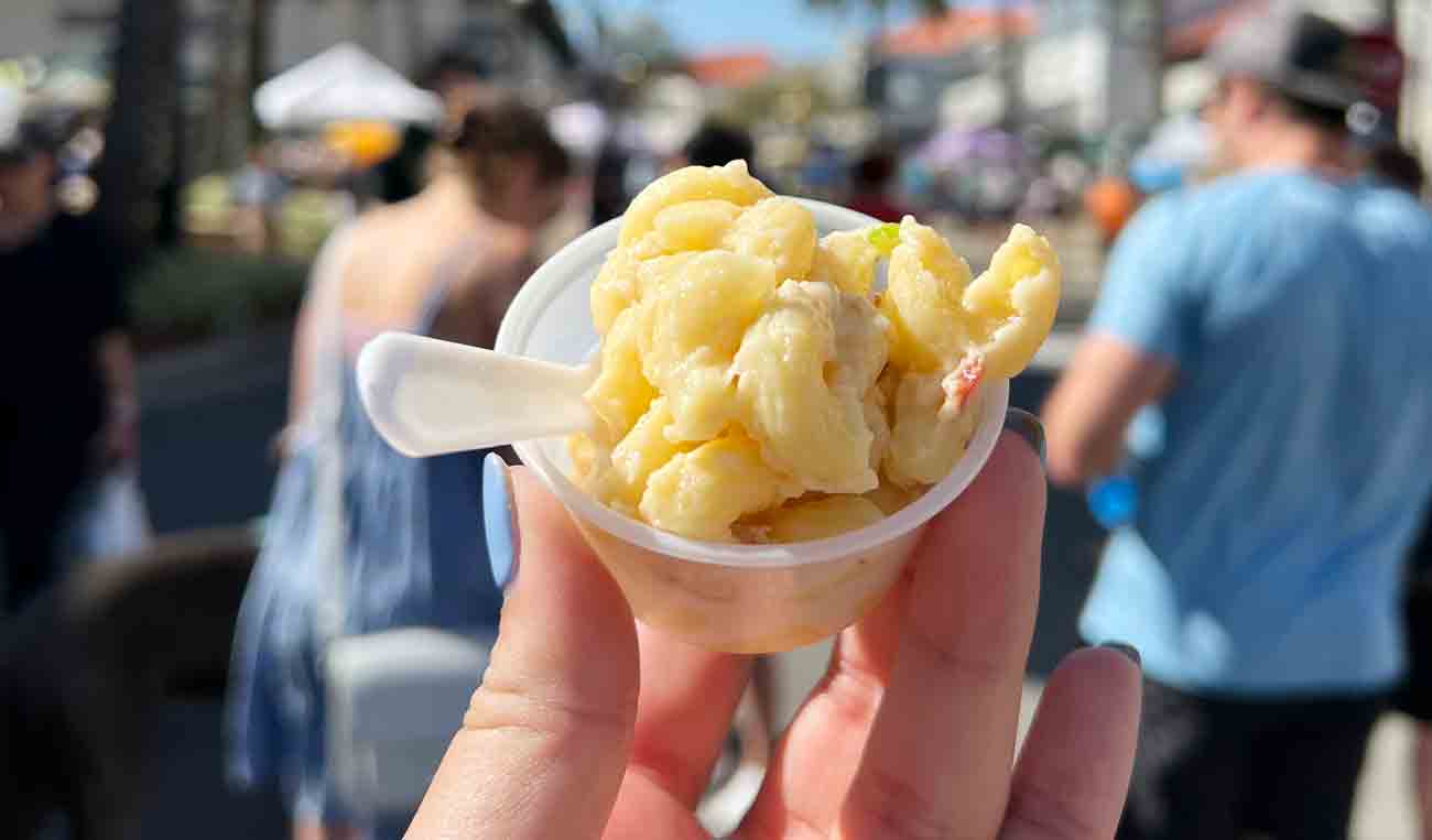 Close-up of a small, hand-held sample of macaroni & cheese (in a plastic container with a plastic fork) with the festival in the background