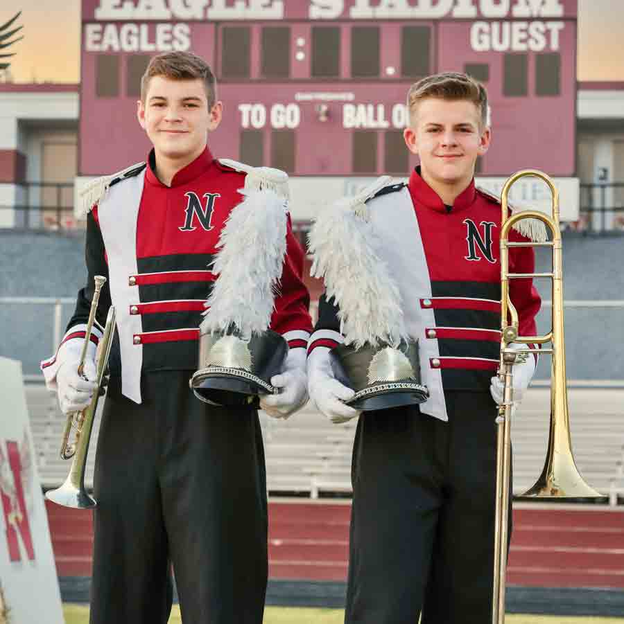 Tommy Roberts and Andrew Roberts, side-by-side in Niceville High School Football stadium with scoreboard behind them. both wearing band uniforms, holding instruments and hats.