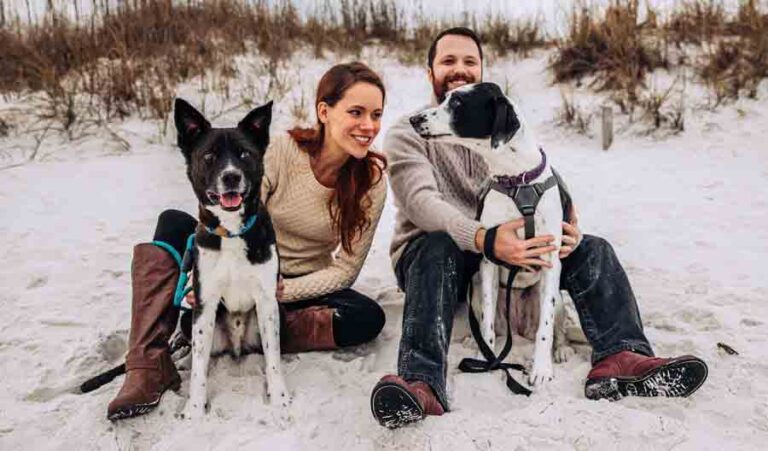 Sara and Joseph Lee are on the sand at the beach with sand dunes and sea oats behind them and each with a dog seated in front of them.