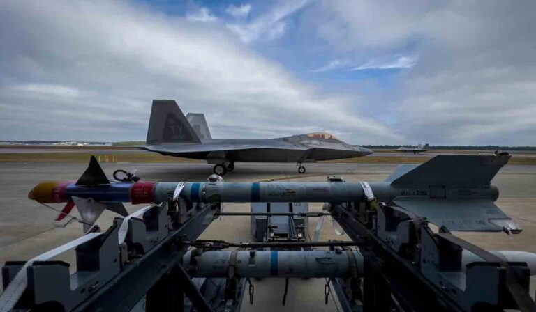 An AIM-9 missile waits on the flightline to be loaded as 325th Fighter Wing F-22A Raptors taxi out for missions in the background.