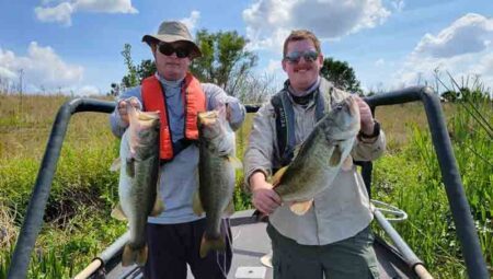 Two biologists are standing onboard a boat holding three trophy-size largemouth bass.