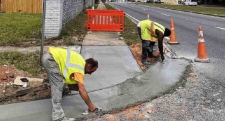 two workers in reflective vests use hand trowels on a newly constructed sidewalk ramp