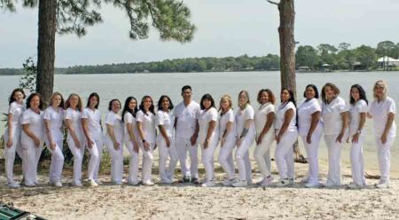 19 adult students in white nursing uniforms in a group pose on a sandy beach with a bayou in the background