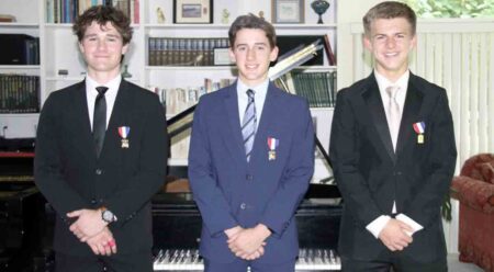 Three teenage young men wearing suits with a gold medal pinned to their coat, standing in front of a piano.