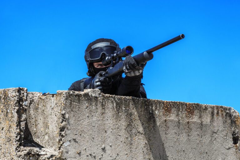 Swat police operator with sniper rifle in black uniform aiming behind concrete block. Sunny day, low angle