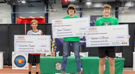 three boys, winners in the state archery tournament, holding oversized checks with a table of trophies in the background