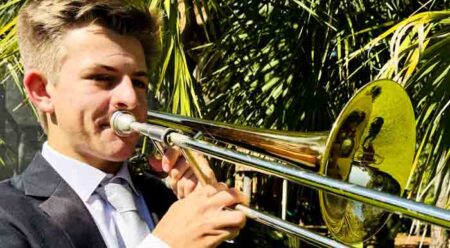 Andrew Roberts playing trombone outdoors with palm tree leaves in the background