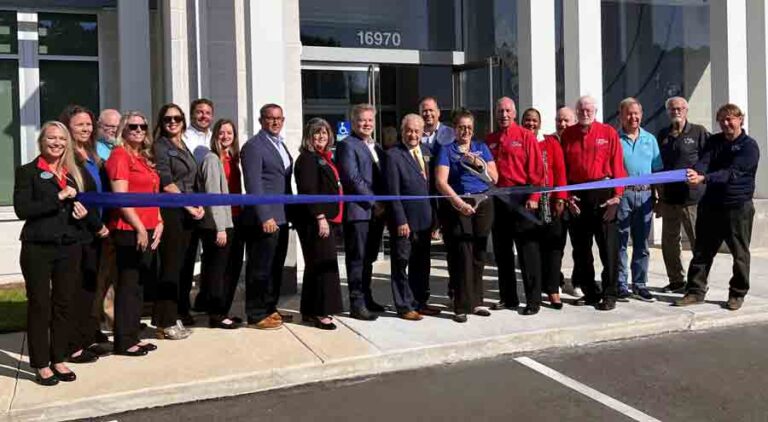 A group of people behind a blue ribbon that is being cut with over-sized scissors.