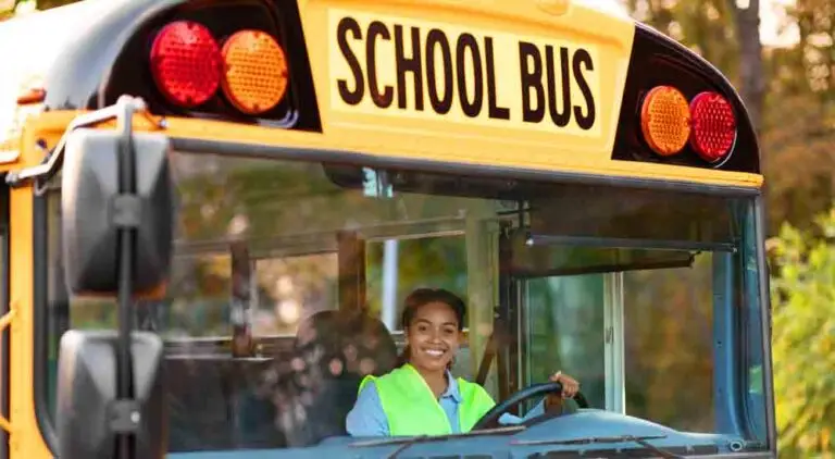 Portrait Of Happy Black Female Driver Driving Yellow School Bus wearing Reflecting Vest Looking Through Window, Holding Steering Wheel And smiling