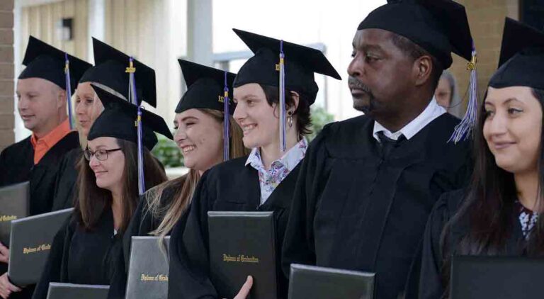 A group of seven men and women graduates wearing black caps and gowns and holding their high school diplomas.