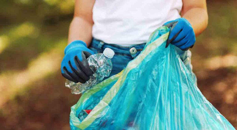 Closeup of gloved hand with a plastic water bottle held over a trash bag during a beach and park cleanup event