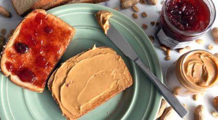 Two slices of bread on a plate with strawberry jam on one slice and peanut butter on the other. A jar of jam and a jar of peanut butter are next to the plate.