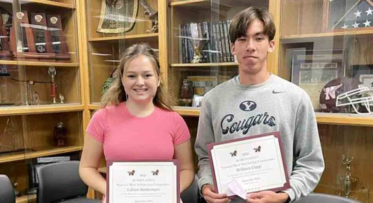 Two high school students, a young woman in a pink T-shirt and blue jeans and a young man in a gray sweater with "Cougars" written on it, stand side by side holding National Merit Scholarship Program certificates. They are in a school setting, surrounded by display cases filled with trophies, awards, and memorabilia.