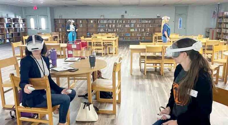 Teachers in a school library wearing VR headsets during a Prism VR training session, with tables and bookshelves in the background.