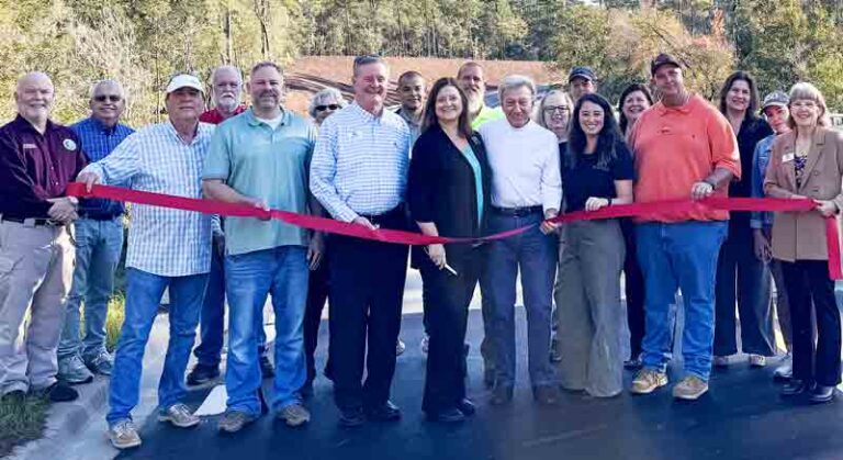 Close-up view of a group of local leaders and community partners standing together during the ribbon-cutting ceremony for the new road and north bridge at Deer Moss Creek in Niceville, Florida, with a backdrop of trees and a sunny sky.