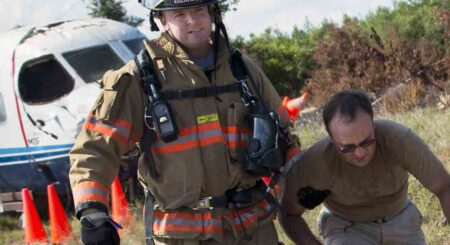 A firefighter assists a man during a simulated aircraft crash response drill near a damaged airplane fuselage in an open field.