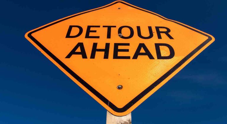 A bright orange diamond-shaped "Detour Ahead" sign mounted on a wooden post against a clear blue sky.