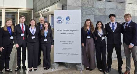 A group of eight Niceville High School students and their chaperones pose in front of the Shizuoka Convention & Arts Center during the 2nd World Congress of Marine Stations in Japan. They are dressed in formal attire, standing next to a sign displaying event details.