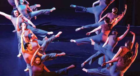 A group of dancers in gray costumes perform a synchronized movement on stage, captured under dramatic stage lighting.