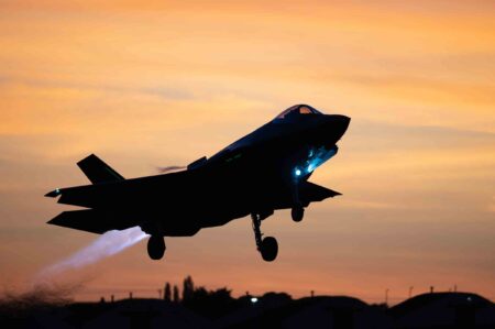 A fighter jet takes off at sunset, with its afterburner glowing and landing gear extended.