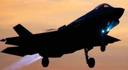 A fighter jet takes off at sunset, with its afterburner glowing and landing gear extended.