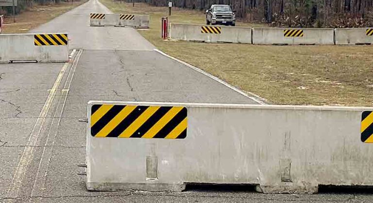 Concrete barriers block Range Road 213 at Eglin Air Force Base, with warning signs indicating restricted access. A vehicle is parked nearby under a cloudy sky, and trees line both sides of the road.