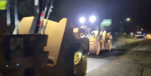 Road resurfacing in progress at night with heavy equipment and bright work lights illuminating the area.