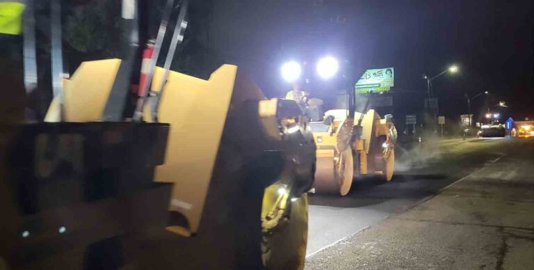 Road resurfacing in progress at night with heavy equipment and bright work lights illuminating the area.