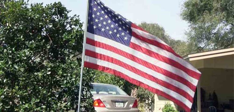 An American flag waves on a metal pole in front of a house, with a car parked in the driveway and greenery in the background.