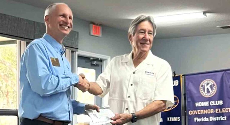 Jeremy Riehl, Kiwanis Florida District Governor, presents a Distinguished Club award to Dr. Bo Arnold, Niceville-Valparaiso Club President, during the Kiwanis Mid-Year Regional Conference in Tallahassee, Florida, on February 1, 2025. The two men are shaking hands while smiling, with Kiwanis banners displayed in the background.