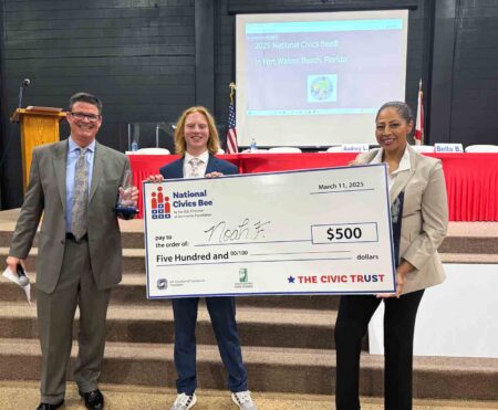 Noah F., the first-place winner of the National Civics Bee in Fort Walton Beach, holds a large check for $500 while standing with event organizers.