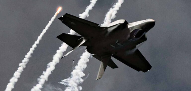 An F-35 fighter jet from Eglin Air Force Base releases flares while flying upside down against a dark sky. White smoke trails follow the aircraft's maneuver.