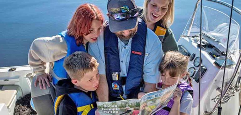 A man wearing a life jacket and hat reads a fishing regulation guide to three children and a woman on a boat.