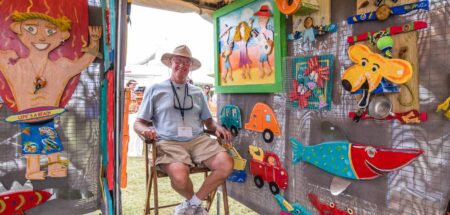 An artist sits in a festival booth surrounded by colorful handcrafted artwork, including wooden fish, beach-themed decor, and whimsical paintings.