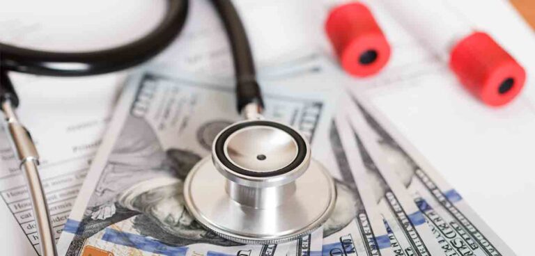 Stethoscope on hundred-dollar bills with medical documents and blood sample vials.