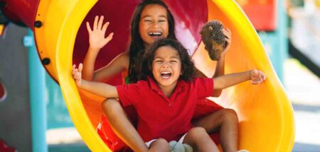 Two smiling children slide down a bright yellow playground slide.