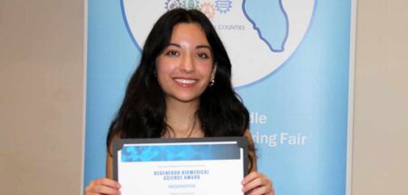 Sasha McCraine holds her Regeneron Biomedical Science Award certificate while standing in front of an East Panhandle Regional Science and Engineering Fair banner.