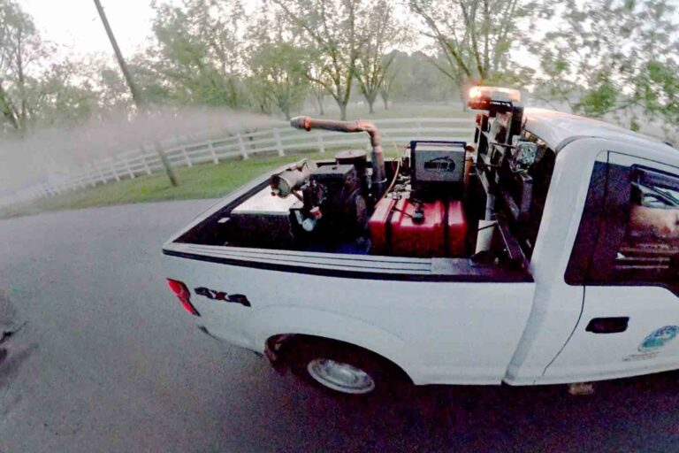 Mosquito control truck spraying pesticide on a neighborhood road at dusk