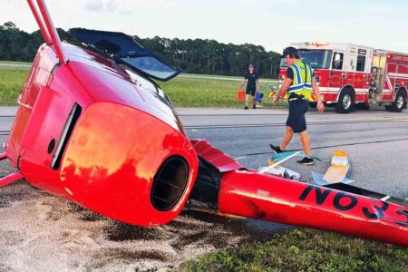 Overturned red helicopter lying on its side at Destin Executive Airport with emergency personnel nearby.