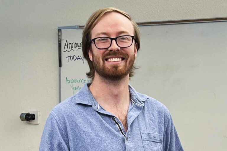 Sean Gordon smiles in a classroom, standing in front of a whiteboard.