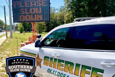Okaloosa County Sheriff's Office patrol vehicle parked beside a "Please Slow Down" sign.