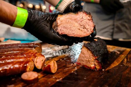 A gloved hand holds a sliced piece of smoked brisket over a cutting board lined with sausages at the 30A BBQ Festival.