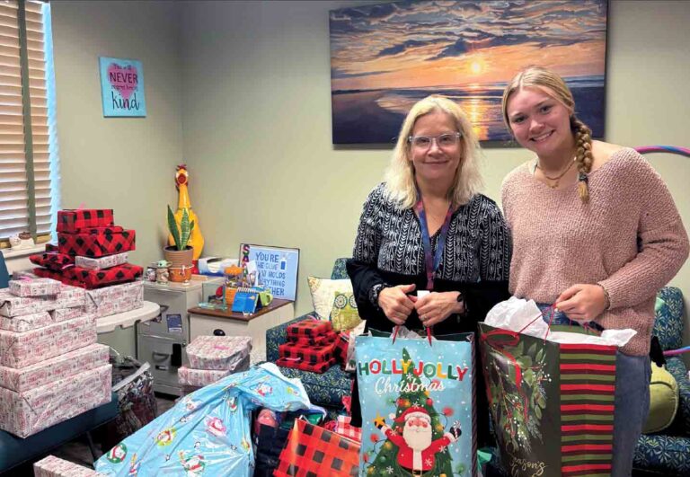 Bailey Howell and Sarah Traver stand together holding gift bags amid stacks of wrapped Christmas presents for the Children in Crisis toy drive.