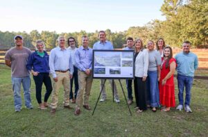 A group of Westonwood Ranch team members, supporters, and community leaders stand outdoors beside a display board showing renderings of the planned life skills and job training center.