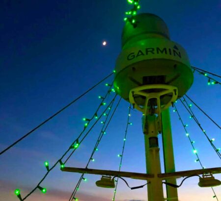 Green Christmas lights wrapped around a boat radar tower at dusk, with the moon visible in the background.