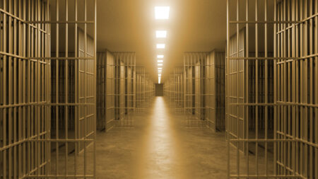 A long, empty prison corridor lined with open jail cells under bright overhead lights.