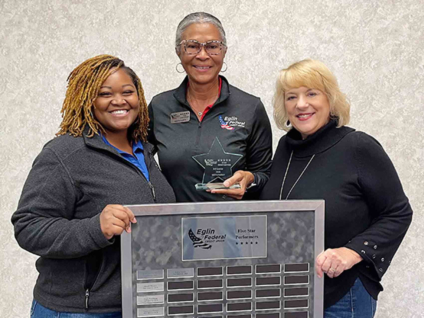 Three Eglin Federal Credit Union employees stand together smiling as Gerta Hardy holds her 5-Star Performer Award beside a recognition plaque displaying past recipients’ names.