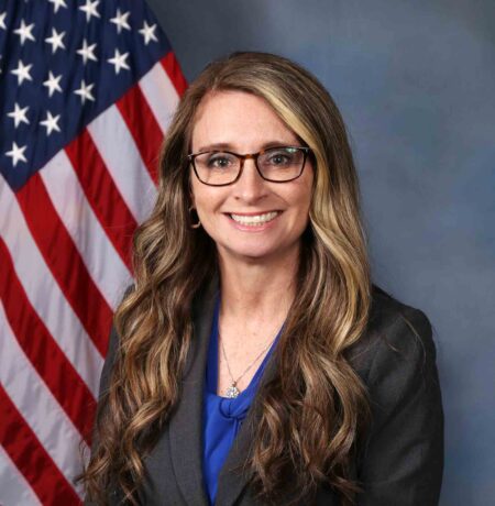 A woman in professional attire is seated in front of an American flag, smiling at the camera.