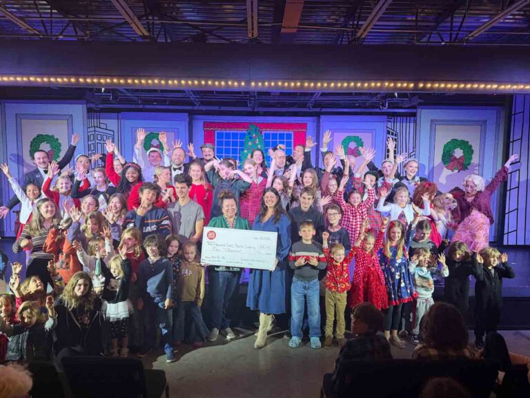 A large group of children and adults on stage celebrate while holding an oversized check awarded to Emerald Coast Theatre Company.