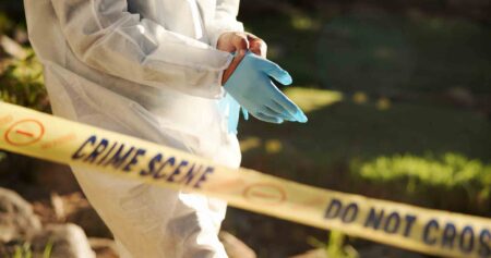 Close-up of a forensic investigator wearing gloves behind yellow crime scene tape outdoors.